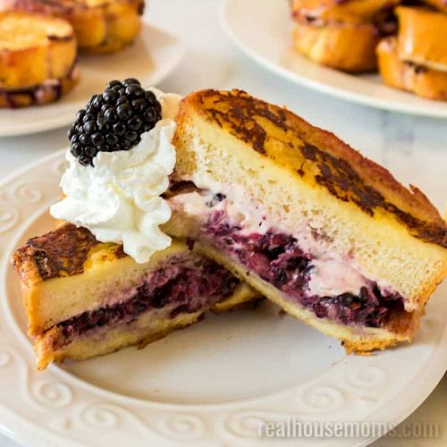 The image shows a plate with a sandwich filled with what looks like blackberries and cream cheese, topped with whipped cream and a blackberry.