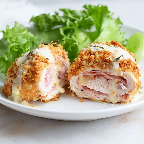 The image shows a plate with sliced, breaded chicken cordon bleu and a side of leafy green lettuce, placed on a white surface.