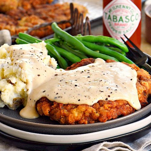 The image shows a plate with chicken-fried steak covered in gravy, mashed potatoes, green beans, and a bottle of Tabasco sauce in the background.