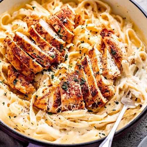 The image shows a creamy pasta dish with sliced, seasoned chicken breast served in a pot with a fork ready to eat.