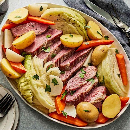 A platter of sliced meat with boiled potatoes, cabbage, and carrots. A sauce bowl, plates, and cutlery are nearby on a table with a gray cloth.