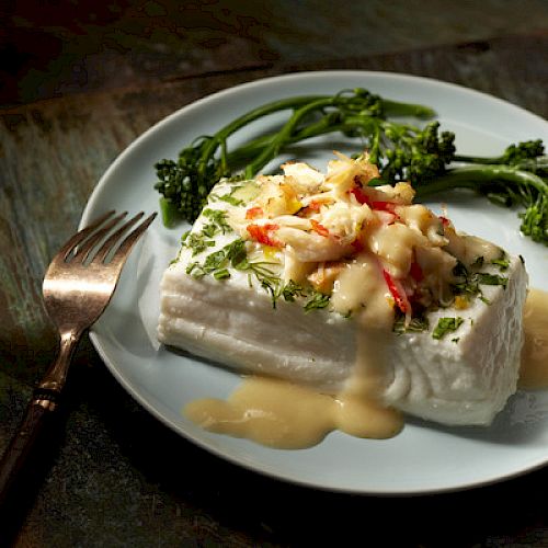 The image shows a plated dish with a piece of white fish topped with a creamy sauce and herbs, accompanied by broccolini, with a fork beside it.