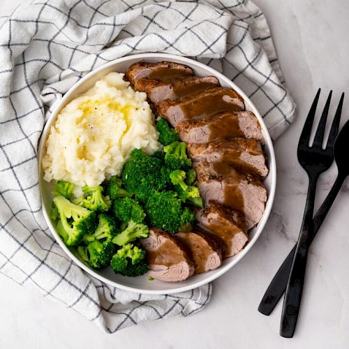 A bowl containing mashed potatoes, steamed broccoli, and sliced meat with sauce on top, placed on a cloth with two black utensils beside it.